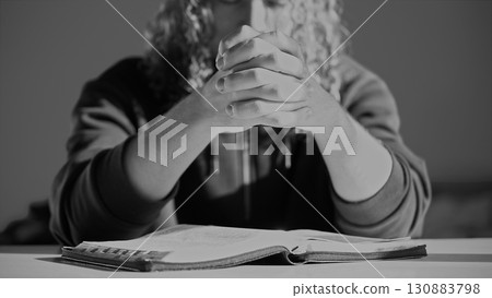 A man with curly hair sits in a worship room, praying intently over a Bible with his palms together. Searching for an answer overcoming a crisis. A scene of faith, solution search, spiritual devotion 130883798