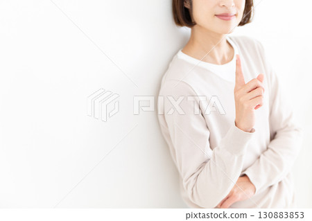 A middle-aged woman in casual clothes leaning against a white wall by the window and pointing up her index finger A middle-aged woman in casual clothes leaning against a white wall by the window and pointing up her index finger 130883853