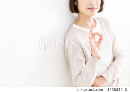 A middle-aged woman in casual clothes leaning against a white wall by the window and making a circle sign 130883940