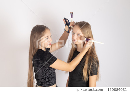 One girl is applying makeup to another girl's face using a brush and a palette. 130884081