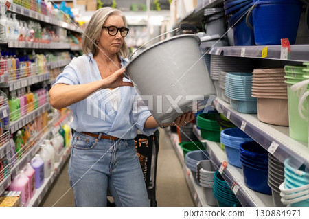 Woman shopping for storage containers in a retail store aisle while selecting a gray bucket 130884971