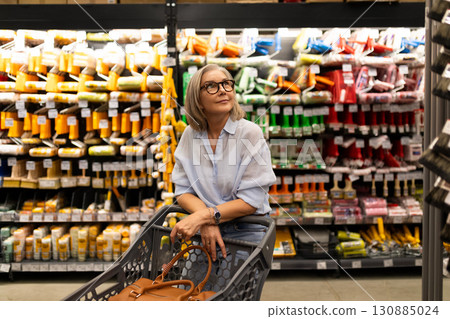Senior woman shopping in a grocery store aisle filled with colorful products while enjoying the 130885024