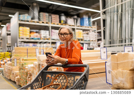 Senior woman shopping for home improvement supplies while using her smartphone inside a warehouse 130885089