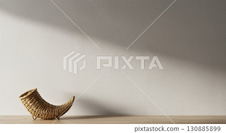 A rustic woven cornucopia rests on a light wooden surface against a plain wall, illuminated by natural light casting a long diagonal shadow, creating a minimalist autumn scene. 130885899