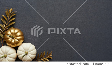 A top-down arrangement of three decorative pumpkins, one gilded gold and two pristine white, nestled with golden foliage on a dark charcoal background, offering ample copy space. 130885908