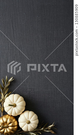 A minimalist top-down arrangement featuring two white and one gold decorative pumpkin with olive branches on a dark textured background, providing ample copy space. 130885909