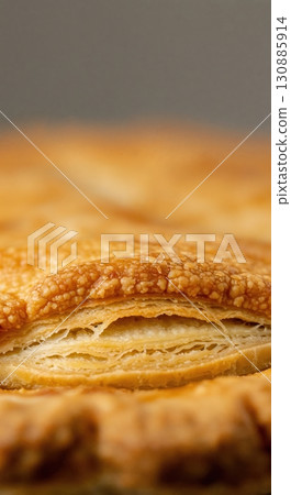 Close-up of a golden, flaky pastry crust with intricate layers and a textured surface, illuminated by soft, warm light against a dark, blurred background. 130885914