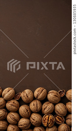 An assortment of whole walnuts and one cracked open nut with its kernel visible, arranged along the bottom edge of a textured dark brown background providing ample copy space. 130885935