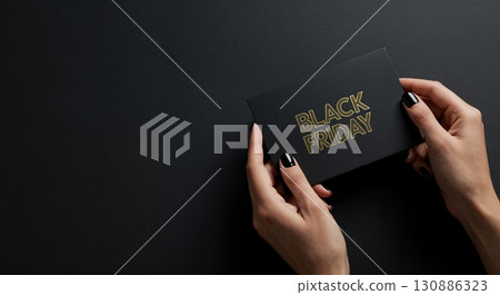 A person's elegant hands with polished dark nails carefully hold a sleek black card featuring the golden "BLACK FRIDAY" text, presented on a minimalist dark surface. 130886323