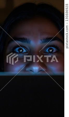 A young woman's wide eyes reflect a shopping cart icon from a screen, illuminated by blue light in a dark setting, showing intense focus or surprise. 130886928