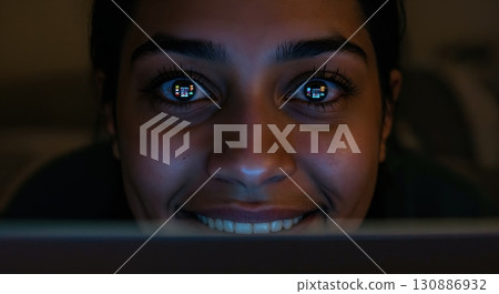 A smiling young adult woman with dark hair is illuminated by the glow of a digital screen, reflecting colorful content in her wide-open eyes within a dark room. 130886932