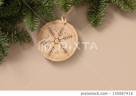 Christmas toy made of decorative wooden slice with ornament snowflake hangs evergreen branches on beige background. Handmade hobby. DIY. Nature material. Xmas decoration. Eco friendly. Zero waste 130887418