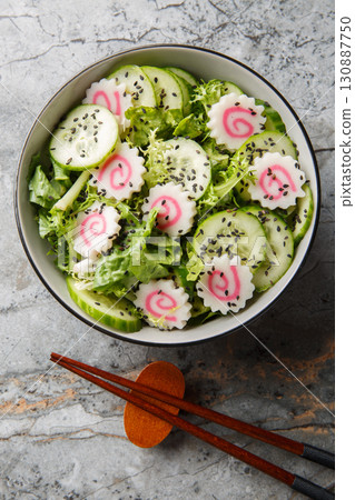 Narutomaki fish roll salad with cucumber, lettuce, green onion and sesame dressing close-up in a bowl on the table. Vertical top view Narutomaki fish roll salad with cucumber, lettuce, green onion and sesame dressing close-up in a bowl on the table. Vertical top view 130887750