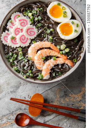 Japanese Buckwheat soba noodles with narutomaki fish roll, shrimp, eggs, sesame seeds close-up in a bowl on the table. Vertical top view Japanese Buckwheat soba noodles with narutomaki fish roll, shrimp, eggs, sesame seeds close-up in a bowl on the table. Vertical top view 130887762