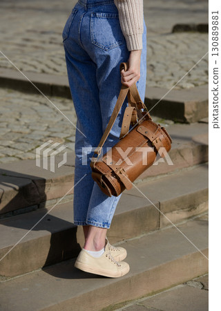 A close-up shot of a model in a knit sweater and jeans, showcasing a unique cylindrical leather bag with intricate side detailing and a belt. A close-up shot of a model in a knit sweater and jeans, showcasing a unique cylindrical leather bag with intricate side detailing and a belt. 130889881