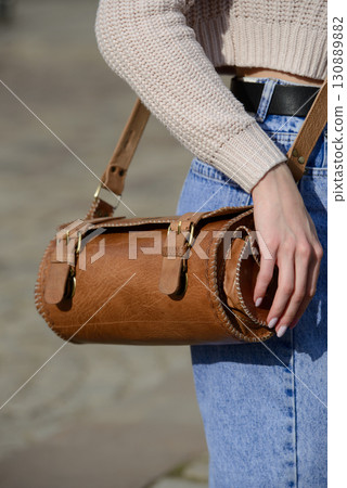 A close-up shot of a model in a knit sweater and jeans, showcasing a unique cylindrical leather bag with intricate side detailing and a belt. A close-up shot of a model in a knit sweater and jeans, showcasing a unique cylindrical leather bag with intricate side detailing and a belt. 130889882