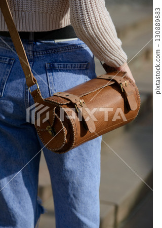 A close-up shot of a model in a knit sweater and jeans, showcasing a unique cylindrical leather bag with intricate side detailing and a belt. A close-up shot of a model in a knit sweater and jeans, showcasing a unique cylindrical leather bag with intricate side detailing and a belt. 130889883