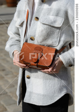 A stylish model in a plaid overshirt holds a small, reddish-brown leather bag with a unique flap and wide buckle, showcasing casual elegance 130889898