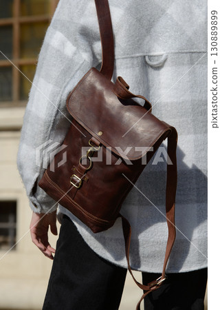 A stylish model is photographed from the back, showcasing a classic brown leather backpack and a trendy plaid overshirt against an urban backdrop A stylish model is photographed from the back, showcasing a classic brown leather backpack and a trendy plaid overshirt against an urban backdrop 130889899