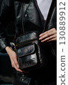 A stylish model in a black leather jacket showcases a unique black leather cross-body bag with a glossy sheen and a front flap pocket 130889912