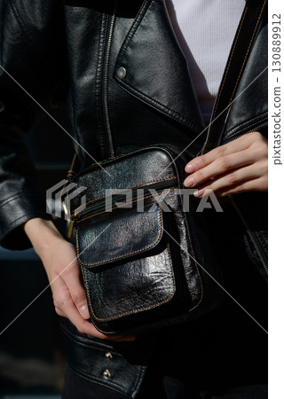 A stylish model in a black leather jacket showcases a unique black leather cross-body bag with a glossy sheen and a front flap pocket 130889912