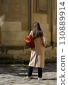 A beautiful fair-skinned model with long, dark hair is photographed from the back, showcasing a sophisticated brown leather backpack and a timeless beige trench coat. 130889914