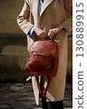 A stylish female model showcasing a brown leather backpack with detailed zippers, held against a sophisticated beige trench coat in an urban setting 130889915