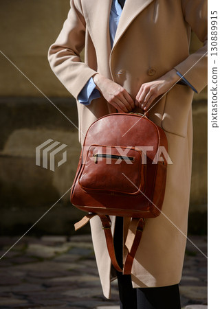 A stylish female model showcasing a brown leather backpack with detailed zippers, held against a sophisticated beige trench coat in an urban setting 130889915
