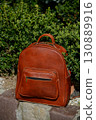 A high-quality brown leather backpack with a front zipper pocket is showcased on a stone ledge in a natural, outdoor setting, highlighting its rugged elegance. 130889916