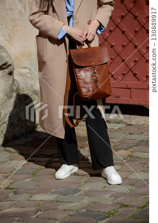 A stylish female model in a classic beige coat and sneakers showcases a sophisticated brown leather backpack with a unique flap closure and buckle detail 130889917