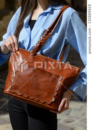 A professional close-up shot of a model showcasing a refined brown leather tote bag with unique braided handles and a decorative front pocket 130889928