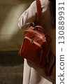 A beautiful fair-skinned model with long, dark hair is photographed from the back, showcasing a sophisticated brown leather backpack and a timeless beige trench coat. 130889931