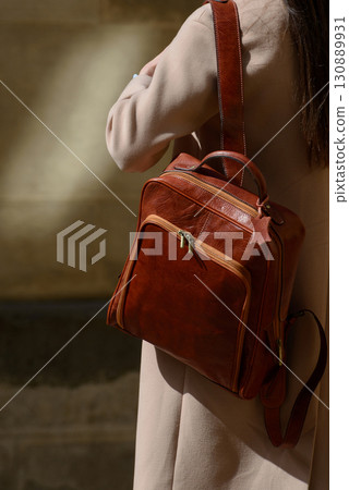A beautiful fair-skinned model with long, dark hair is photographed from the back, showcasing a sophisticated brown leather backpack and a timeless beige trench coat. A beautiful fair-skinned model with long, dark hair is photographed from the back, showcasing a sophisticated brown leather backpack and a timeless beige trench coat. 130889931