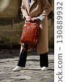 A stylish female model showcasing a brown leather backpack with detailed zippers, held against a sophisticated beige trench coat in an urban setting 130889932