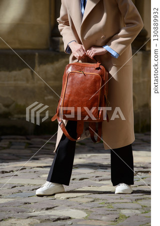 A stylish female model showcasing a brown leather backpack with detailed zippers, held against a sophisticated beige trench coat in an urban setting A stylish female model showcasing a brown leather backpack with detailed zippers, held against a sophisticated beige trench coat in an urban setting 130889932
