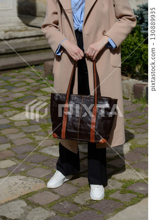 A stylish urban shot captures a woman in a beige coat and sneakers, showcasing a spacious leather tote bag with contrasting handles 130889935