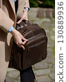 A stylish female model showcasing a brown leather backpack with detailed zippers, held against a sophisticated beige trench coat in an urban setting 130889936