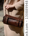 A professional fashion shot capturing a female model showcasing a cylindrical brown leather bag with detailed buckles and a strap, highlighting her beige coat. 130889948