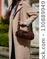 A professional fashion shot capturing a female model showcasing a cylindrical brown leather bag with detailed buckles and a strap, highlighting her beige coat. 130889949