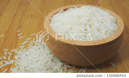 Uncooked white rice grains in a wooden bowl with rice scattered on wooden table, natural food ingredient for Asian cuisine. 130890454