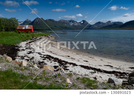 White Sand Beach With Red Hut And Snowy Mountains At Fjord On Lofoten In Norway 130891023