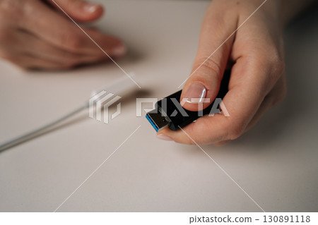 Female hand holding black USB flash drive with blue connector, positioned near white USB adapter on white table, representing digital data storage, close-up. Concept of modern technology. 130891118