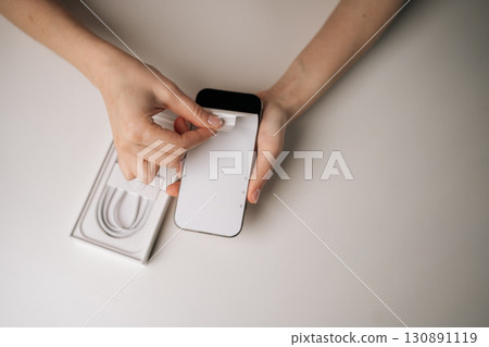 Close-up top view of woman unpacking new smartphone and removing screen protector sitting on table, with charging cable in box visible in background. Concept of excitement of unboxing new device 130891119