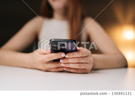 Close-up of female hands holding smartphone, browsing internet, typing messages and using mobile apps in cozy home environment with warm bokeh background. Concept of modern technology. 130891131