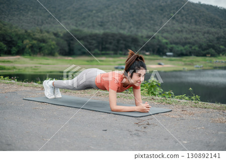 Woman in athletic wear doing a plank exercise on a yoga mat by a lake with green hills. Woman in athletic wear doing a plank exercise on a yoga mat by a lake with green hills. 130892141