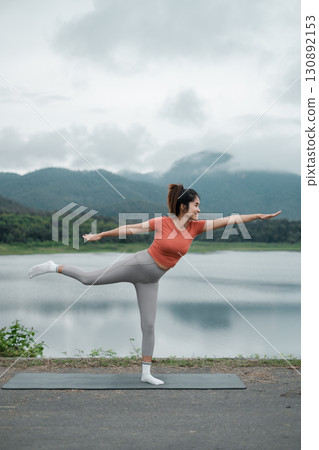 A woman performs a yoga pose on a mat by a tranquil lake with mountains in the background, under a cloudy sky. 130892153