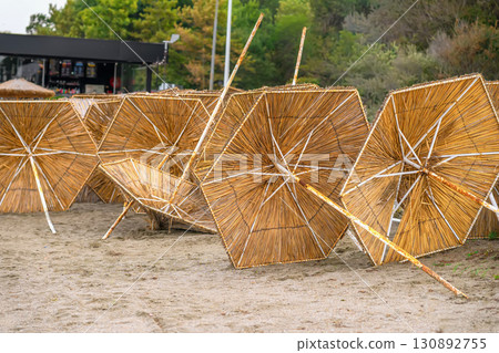 Straw Beach Umbrellas. A row of straw beach umbrellas lying on sandy ground near a beach. Concept of seaside relaxation, end of summer, end of holiday season Straw Beach Umbrellas. A row of straw beach umbrellas lying on sandy ground near a beach. Concept of seaside relaxation, end of summer, end of holiday season 130892755