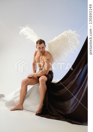 Male model sits gracefully with white wings and a dark drape in studio setting 130893149
