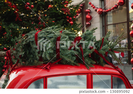 Christmas tree transported on red car with holiday decoration. Festive holiday scene at modern city. Concept of delivering goods. Christmas tree transported on red car with holiday decoration. Festive holiday scene at modern city. Concept of delivering goods. 130895092