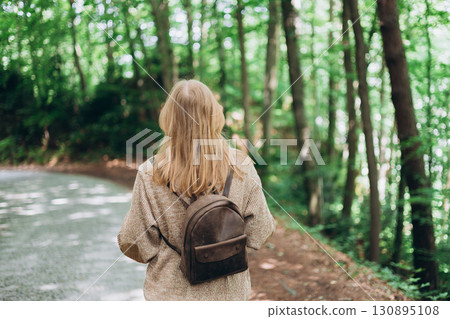 Young adult woman staring at green trees. Thinking about life at beautiful park in spring day. Spending time alone in nature. Peaceful atmosphere. Back view. 130895108